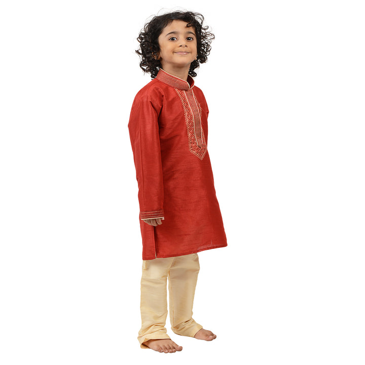 Boys Kurta Set in red art silk with gold embroidered neckline, paired with cream churidar for traditional events and family gatherings.